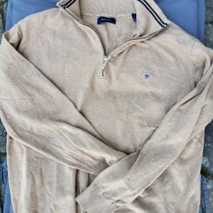 Beige half zip tröja från Gant - Snygg beige tröja från Gant med half zip och ribbad krage med mörkblå ränder. Tröjan har lång ärm och broderad Gant-logga på bröstet. Perfekt för lager-på-lager och chill stil. Säljer pågrund av att den är lite fö stor. Doftfri. 