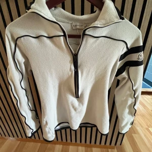 Vit fleece half zip från Moncler - Vit fleece-tröja från Moncler med svarta detaljer och broderad logga på ärmen. Tröjan har half zip, lång ärm och svarta ränder på ena ärmen. Perfekt för kyliga dagar och riktigt snygg till jeans eller tights.