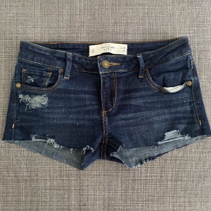Lågmidjade jeansshorts - Snygga mörkblå jeansshorts från Abercrombie & Fitch med slitna detaljer och råa kanter. Midjemått: 37 cm rakt över, grenhöjd: 17 cm.
