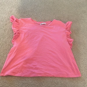 Rosa volangtopp från Zara - Supersöt rosa topp från Zara med volangdetaljer längs axlarna. Modellen är ärmlös och har en rund halsringning. Perfekt för dig som gillar färg och vill ha något som sticker ut lite extra. Tillverkad i mjuk bomull för en skön känsla.
