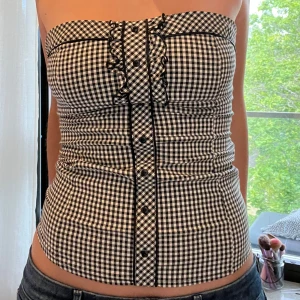 Rutig tubtopp från Mohito - Supersnygg svartvit rutig tubtopp från Mohito med knappar framtill och volangdetalj längs knappraden. Toppen är tight och har bandeauform, perfekt till jeans eller kjol. Tunt och bekvämt material som känns lätt på kroppen.