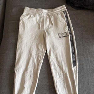 Ljusgråa mjukisbyxor från Emporio Armani - Snygga ljusgråa mjukisbyxor från Emporio Armani med svarta sidoremsor och logga längs benen. Byxorna har dragsko i midjan och EA7-logga framtill. Perfekta för en chill och stilren look. Dom har en mindre reva på sidan men inget som syns mycket där av den billigare priset.Size 158