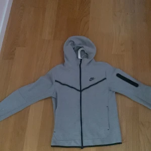Grå Nike hoodiejacka med dragkedja S - Säljer en grå hoodiejacka från Nike i storlek S. Jackan har hel dragkedja, huva och svarta detaljer längs bröstet och på ärmen. Perfekt för chill dagar eller när du vill ha en sportig look. Mjuk och skön med två fickor framtill.