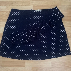 Mörkblå prickig kjol från Springfield - Supersöt mörkblå kortkjol från Springfield med vita prickar och volangdetalj framtill. Kjolen har en klassisk A-linjeform och är perfekt för dig som gillar en lekfull och trendig stil. Lätt och skön att bära, passar perfekt till sneakers eller sandaler. 