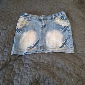 Miss Chic jeanskjol med broderi - Snygg ljusblå jeanskjol från Miss Chic med slitna detaljer och coola broderier i vitt och grönt på fickorna. Kjolen har klassisk femficksdesign, bälteshällor och dragkedja framtill. Limited Edition-märke baktill ger extra edge till looken. Storlek 38/27.