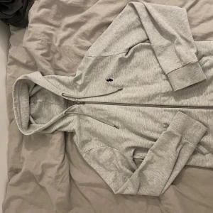 Grå hoodiejacka från Polo Ralph Lauren - Ljusgrå hoodiejacka från Polo Ralph Lauren med dragkedja framtill, justerbar huva med snören och klassisk broderad logga på bröstet. Tillverkad i mjuk bomullsmix, perfekt för chill dagar. Ribbade muddar vid ärmslut och nederkant för extra komfort.