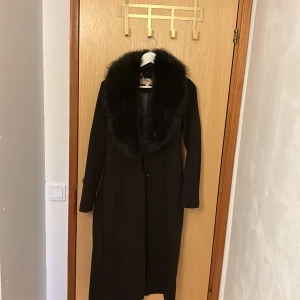Brun  kappa med fuskpäls från Michael Kors - Lång brun kappa från Michael Kors med bred krage i fuskpäls och snyggt bälte i midjan. Klassisk och elegant look med guldiga detaljer på insidan. Perfekt för dig som vill ha en stilren vibe under kallare dagar.