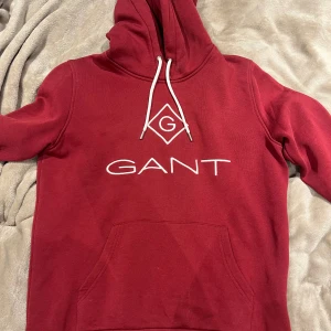 Vin röd gant hoodie - Röd hoodie från GANT med vit broderad logga och grafisk detalj på bröstet. Tröjan har huva med vita snören och en stor känguruficka framtill. Tillverkad i mjuk bomull, passformen är mindre i storleken så passar xs-s använd sparsamt inga defekter pris 199