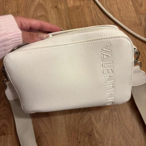 Vit axelväska från Valentino - Snygg vit axelväska från Valentino med präglad logga på framsidan och bred, justerbar axelrem med tryckt logotyp. Väskan har två fack med dragkedja och är gjord i strukturerat skinnliknande material. Perfekt för att lyfta din outfit.