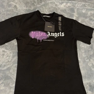 Palm Angels svart t-shirt Shanghai - Svart t-shirt från Palm Angels med tryck i vitt och lila på bröstet där det står 'Palm Angels Shanghai'. T-shirten har klassisk passform, rund hals och korta ärmar. Materialet är mjuk bomull och trycket har en cool spray paint-effekt.