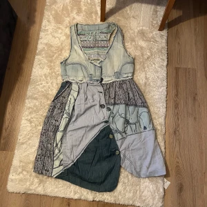 Klänning i jeans och bomull - Unik ärmlös klänning i ljusblå denim och olika blå och grå bomullstyger. Klänningen har broderade blommor, dekorativa knappar framtill och råa kanter. Snyggt mönstrad insida och cool boho-vibe. Perfekt för dig som gillar en kreativ och avslappnad stil.