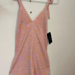 Randig kortklänning från NA-KD - Superfin stickad kortklänning från NA-KD i pastelliga rosa, lila och beige toner. Klänningen har smala axelband med knyt, v-ringning och ett randigt melerat mönster, den är öppen i ryggen med en knytning runt nacken. För kort för mig men kan skicka bild på om man vill. Helt oanvänd. 