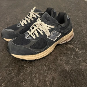 New Balance 2002R mörkgrå sneakers - Snygga New Balance 2002R sneakers i mörkgrå mocka och mesh med beige sula. Klassisk siluett med vita snören och reflekterande detaljer. Perfekta för dig som gillar streetwear och vill ha en bekväm och stilren sko till vardags.
