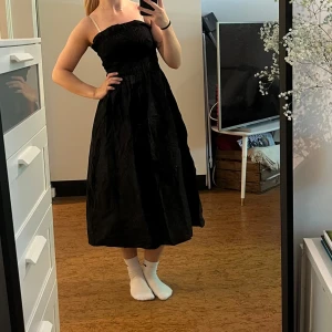 Svart långklänning med smock - Svart långklänning med smockad topp och axelbandslös design. Klänningen har en luftig, vid kjol och volangdetalj upptill. Materialet känns lätt och passar bra till varmare dagar. Helt oanvänd