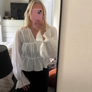 Vit blus med v-ringning och volang - Superfin vit blus i tunn, luftig bomull med v-ringning och långa ärmar. Blusen har lätt rynkade detaljer och volang nedtill som ger en romantisk vibe. Perfekt till jeans eller kjol för en fräsch look. Helt ny aldrig använd! Skriv för mer bilder 