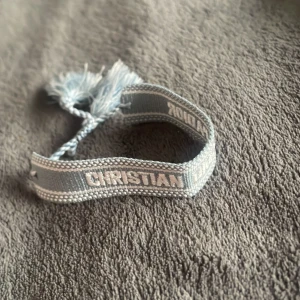 Blått Christian Dior J'adior armband - Snyggt vävt armband från Christian Dior i ljusblått med texten 'Christian Dior J'adior' broderad i vitt. Armbandet har justerbar knytning med tofsar i ändarna för en cool och avslappnad look. Perfekt accessoar för att lyfta din outfit.