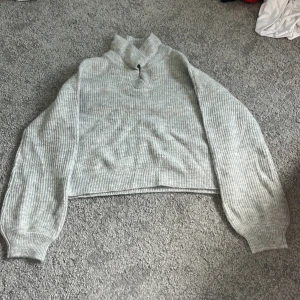 Grå ribbad polotröja från Kappahl - Mysig grå polotröja från Kappahl i ribbstickat material. Tröjan har en hög krage och långa ärmar med breda muddar. Perfekt för kyliga dagar och enkel att matcha med jeans eller kjol.