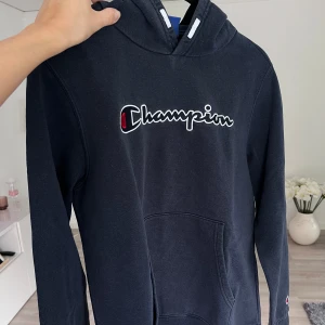 Mörkblå Champion-hoodie - Säljer en mörkblå hoodie från Champion i storlek 2XL. Tröjan har klassisk huva med vita snören, stor magficka och broderad Champion-logga på bröstet och ärmen. Perfekt för dig som gillar streetwear och vill ha en skön och stilren look. 💙 Önskar du veta exakta mått? Tveka inte att höra av dig!