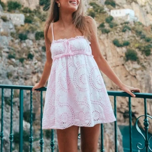 Söt vit och rosa kortklänning - Superfin kortklänning med ljusrosa detaljer och broderade mönster. Klänningen har smala axelband, volang upptill och en söt rosett framtill. Perfekt för varma sommardagar och ger en romantisk vibe.