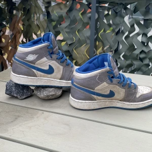 Nike Air Jordan 1 grå/blå sneakers - Nike Air Jordan 1 sneakers i grått och vitt med blå detaljer och blå swoosh. Skorna har högt skaft, blå snörning och klassisk Air Jordan-logga på sidan. Ovandelen är i skinn och textil, med perforerad tå för extra ventilation.