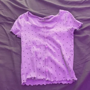 Vit prickig topp från H&M - Supersöt vit topp från H&M med svarta prickar, korta ärmar och knappar framtill. Ribbad struktur och volangkant vid ärmslut och nederkant ger en gullig vibe. Perfekt för dig som gillar pasteller och lekfulla detaljer.