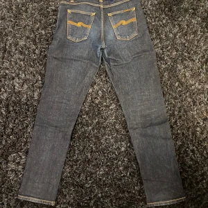 Mörkblå Nudie jeans - Snygga mörkblå jeans från Nudie med klassiska orange sömmar och broderade bakfickor. Modellen har fem fickor, raka ben och normal passform. Tillverkade i slitstarkt jeanstyg med guldfärgade detaljer och knappstängning framtill.