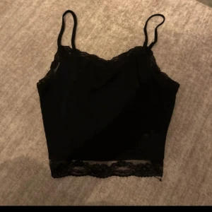 Svart ribbad linnetopp med spets - Snygg svart croppad linnetopp med tunna axelband och ribbad struktur. Toppen har fina spetsdetaljer längs halsringning och nederkant som ger en extra touch. Perfekt att styla med jeans eller kjol för en trendig look.