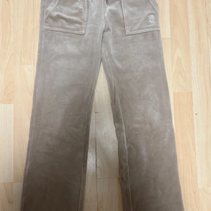 Beige velourbyxor Juicy Couture XXS - Supermjuka beige mjukisbyxor från Juicy Couture i velour med breda ben och stora fickor framtill. Byxorna har snörning i midjan och broderad JC-logga på ena fickan. Perfekta för en chill och trendig look.