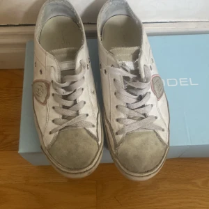 Vita Philippe Model sneakers med glitterdetalj - Snygga vita Philippe Model sneakers med grå detaljer och glittrig sköld på sidan. Klassisk låg modell med snörning och platt sula. Skorna har en cool, urban vibe och är tillverkade i skinn med inslag av mocka på tån och hälen.