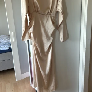Lång beige klänning med vida ärmar - Säljer en elegant långklänning i beige med vida, långa ärmar och markerad midja. Klänningen har en mjuk, slät yta och en lätt glans som ger en lyxig känsla. Perfekt för dig som gillar minimalistisk och stilren design.