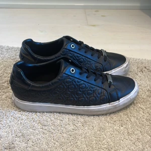 Svarta sneakers från Calvin Klein - Snygga svarta sneakers från Calvin Klein med präglad CK-logga över hela skon. Skorna har vita sulor, svarta skosnören och detaljer i metall framtill. Tillverkade i skinn för en clean och stilren look som passar till det mesta.