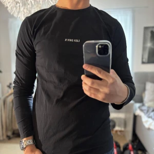 Svart långärmad tröja Les Deux - Svart långärmad tröja från Les Deux med vit broderad logga på bröstet. Tröjan har rund halsringning och är gjord i mjuk bomull, perfekt för en clean och stilren look. Passar dig som gillar minimalistisk design och snygga basplagg.