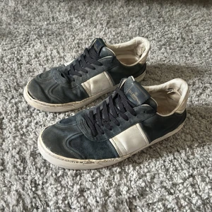 Valentino Flycrews - Snygga Valentino Garavani sneakers i svart mocka och vitt skinn med svarta snören och coola nitar på hälen. Klassisk låg modell med rund tå och vit sula. Perfekta för dig som gillar exklusiva streetwear-vibbar. Mörkblå/Vit färg på skorna. Ganska använda och finns en del defekter men syns inte så mycket vid användning. Går fortfarande att använda utan problem. Kontakta mig om du har frågor.