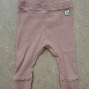 Leggings - Supersöta ljusrosa ribbade leggings från Lindex Baby i mjuk bomullsblandning med elastan. Byxorna har breda muddar vid bensluten och en liten tygpatch med regnbåge framtill. Perfekta för små bebisar som vill ha det bekvämt och stilrent.