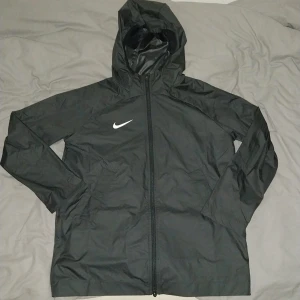 Svart Nike Storm-FIT Windbreaker - Svart windbreaker från Nike med Storm-FIT-teknologi och huva. Jackan har hel dragkedja framtill, lång ärm och klassisk Nike-logga på bröstet. Perfekt för blåsiga dagar och sportiga outfits. Materialet är lätt och vattenavvisande syntet.