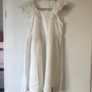 Vit spetsklänning från Zara - Superfin vit klänning från Zara med broderad spets och volangdetaljer på axlarna. Klänningen har en luftig passform och är ärmlös, perfekt för sommarens varma dagar. Söt och romantisk look med vackra detaljer.