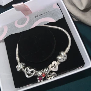 Pandora armband med berlocker - Snyggt silverfärgat armband från Pandora med flera berlocker, bland annat hjärtan och en söt björn med röd och rosa detalj. Armbandet har klassisk ormkedja och glittriga detaljer på vissa berlocker. Perfekt accessoar för att lyfta din stil. Påsen finns med om köparen vill ha det 