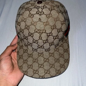 GG keps med GG-mönster och läderdetalj - Snygg keps från GG beige och brunt med klassiskt GG-monogram över hela kepsen. Har grönt och rött band på sidan och justerbar rem i brunt läder baktill. Tillverkad i bomullsmix och detaljer i läder, perfekt för dig som vill sticka ut.