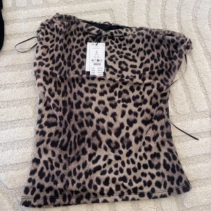 Leopardmönstrad tubtopp från Gina Tricot - Snygg tubtopp från Gina Tricot med leopardmönster i beige och svart. Toppen är axelbandslös och har en tight passform som framhäver siluetten. Perfekt för dig som vill sticka ut med ett djurmönstrat plagg.