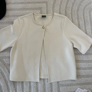 Beige cardigan jacka från gina - Stilren beige cardigan jacka från gina med kort ärm och rund halsringning. Jackan har en enkel knäppning med en dekorativ knapp upptill och är tillverkad i ett mjukt material som ger en clean look.