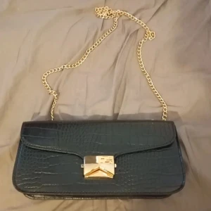 Svart axelväska med kedja - Snygg svart axelväska med krokopräglat mönster och guldfärgad kedjerem. Väskan har ett stilrent, rektangulärt format och ett elegant guldfärgat spänne framtill. Perfekt för dig som vill ha en trendig och klassisk accessoar.