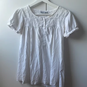 Vit blus med spets från Lindex - Söt vit blus från Lindex med korta puffärmar, knappar framtill och fina spetsdetaljer upptill. Blusen har en lös passform och smockad överdel som ger en romantisk vibe. Perfekt till jeans eller kjol för en fräsch look.