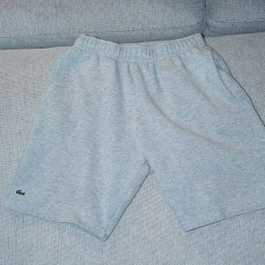 Grå mjuka shorts från Lacoste - Snygga grå shorts från Lacoste med elastisk midja och sidofickor. Klassisk design med den ikoniska gröna krokodilen broderad längst ner. Perfekta för chill dagar eller sport. Tillverkade i mjuk bomull för maximal komfort.