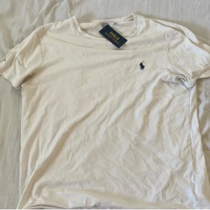 Vit t-shirt från Polo Ralph Lauren - Vit t-shirt från Polo Ralph Lauren med klassisk blå broderad logga på bröstet. Slim fit-modell i mjuk bomull, rund hals och korta ärmar. Perfekt basic-plagg med stilren design och skön passform.