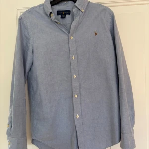 Blå skjorta från Ralph Lauren - Klassisk ljusblå skjorta från Ralph Lauren med button-down krage och broderad logga på bröstet. Skjortan har lång ärm, vita knappar och är tillverkad i bomull. Använd ett fåtal gånger och därför i väldigt bra skick. Hör av er vid frågor :)