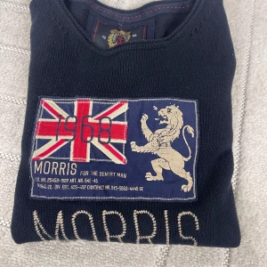 Mörkblå stickad tröja från Morris - Snygg mörkblå stickad tröja från Morris med broderad logga och Union Jack-flagga framtill. Tröjan har lång ärm, rund hals och coolt lejonmotiv. Perfekt för dig som gillar klassisk brittisk stil och vill sticka ut lite extra.