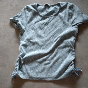 Ljusblå ribbad topp med snörning - Superfin ljusblå ribbad topp från C i storlek 158/164. T-shirten har korta ärmar, rund halsringning och snygga snörningsdetaljer i sidorna som ger en cool look. Perfekt för dig som gillar en enkel men trendig stil.