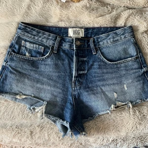 Blå jeansshorts från BDG Urban Outfitters - Snygga blå jeansshorts från BDG Urban Outfitters med råa, fransiga kanter och slitna detaljer. Klassisk femficksmodell med knapp och dragkedja framtill. Perfekta för sommaren och ger en avslappnad vibe. Passar S-L beroende på hur man vill att dem ska passa!