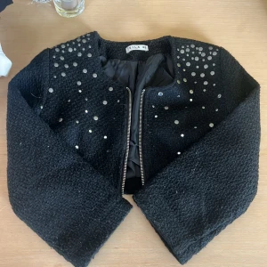Svart cardigan jacka med nitar från Vila - Svart cardigan jacka från Vila i storlek XS. Jackan har en grovstickad struktur, dragkedja framtill och är dekorerad med silvriga nitar på axlar och bröst. Perfekt för dig som vill ha en cool och snygg look.