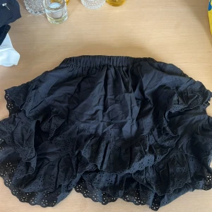 Svart volangkjol med broderade spetsdetaljer - Supersöt svart kortkjol från H&M med flera lager volanger och broderade spetsdetaljer längs kanterna. Kjolen har resår i midjan för en bekväm passform och är perfekt för dig som gillar en feminin och lekfull stil.